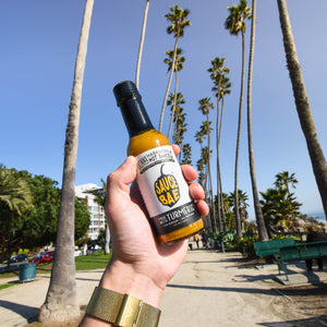 A bottle of Sauce Bae skinny habanero hot sauce being held in the sky with palm trees in the background