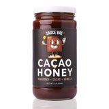 Front view of a 1-pound jar of Sauce Bae Cacao Honey, featuring a cartoon chocolate character on a black label with the text “Cacao Honey – Raw Honey + Cacao + Vanilla.