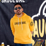 A man wearing a Sauce Bae Hot Sauce Sweatshirt Hoodie in gold. Text logo on front, pepper logo on left sleeve.