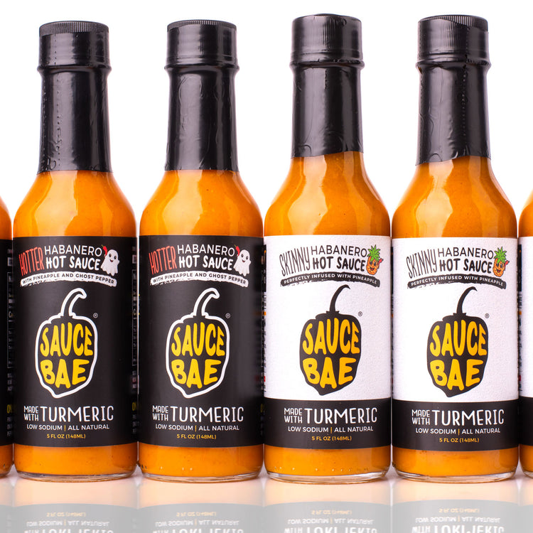 Lineup of Sauce Bae hot sauces featuring alternating bottles of Hotter Habanero with black labels and Skinny Habanero with white labels. Each 5 oz bottle displays the Sauce Bae logo and “Made with Turmeric” tagline. The hot sauces are arranged in a clean row against a white background with subtle reflections below, showcasing the visual contrast between the two flavors.