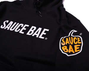 Close up view of front of Sauce Bae Hot Sauce Sweatshirt Hoodie in Black with Sauce Bae Text logo on front, and also pepper logo on sleeve.